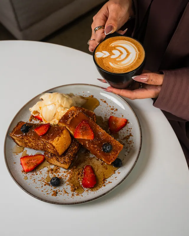 A guest lifting a latte beside French toast with berries and cream